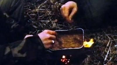 Hand of a soldier stirring a portion of food being cooked over a campfire outdoors.