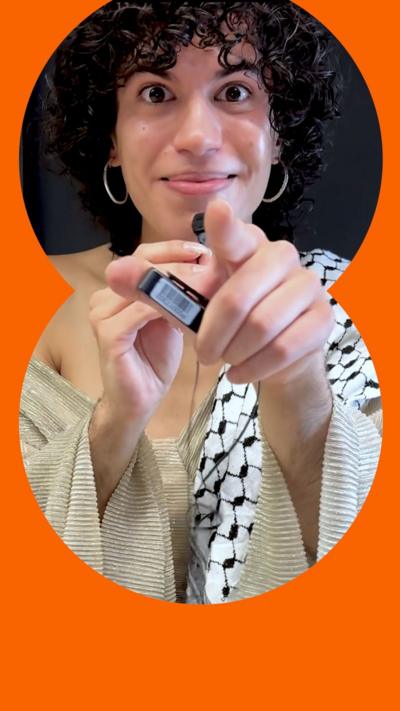 Jacob Alon points at the camera, wearing a flowy silver top. They have curly black hair and silver earrings 