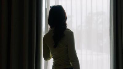 A woman looking out of a window from behind a sheer blind