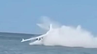 Still of a white personal aircraft crash landing into water. Jet spray is flying up around the plane.
