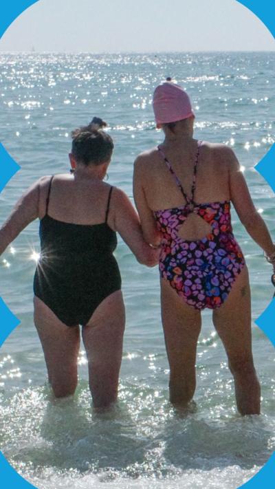 Two women shown from behind holding hands and wading into the sea. The one on the left is wearing a plain black swimsuit. The one on the right is wearing a purple patterned swimsuit and a pale pink swimming cap.