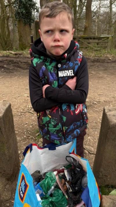 A young boy with an annoyed face and a bag of litter. He is wearing a black jacket with multi coloured patches on the front