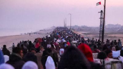 A long crowd of people heads towards the horizon next to a beach in Gaza, a Palestinian flag is flying above