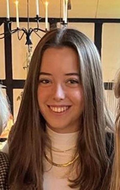 A girl with shoulder length brown hair smiles towards the camera. She is wearing a cream top with a gold necklace