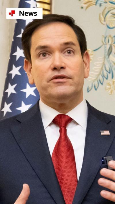 Marco Rubio wearing a blue suit and red tie, standing in front of the US flag