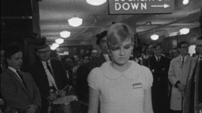 Black and white image of a woman dancing to music played by a piper behind.  She is wearing a name badge (as if she may be a member of staff).  Behinf in the shop, men gaze at her while she dances.  The word 'down' can be seen with an arrow beside it on an overhead sign.