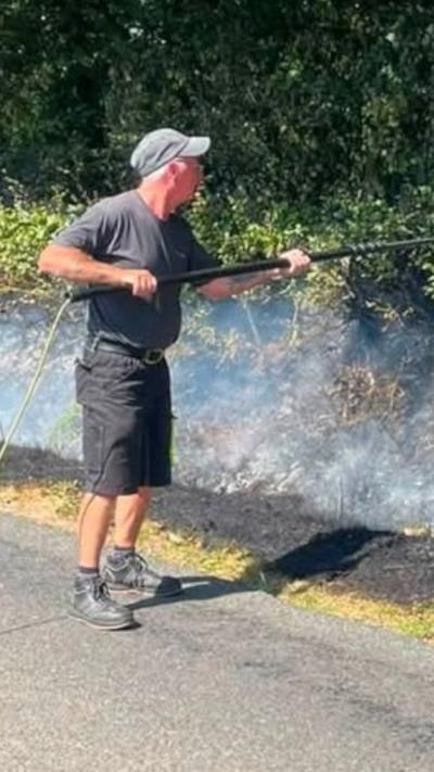 A window cleaner wearing a black T-shirt, shorts and boots uses a window brush to put out a fire on a grass verge at the side of a road