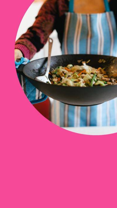 A wok containing stir fry is held by a person wearing a blue striped apron