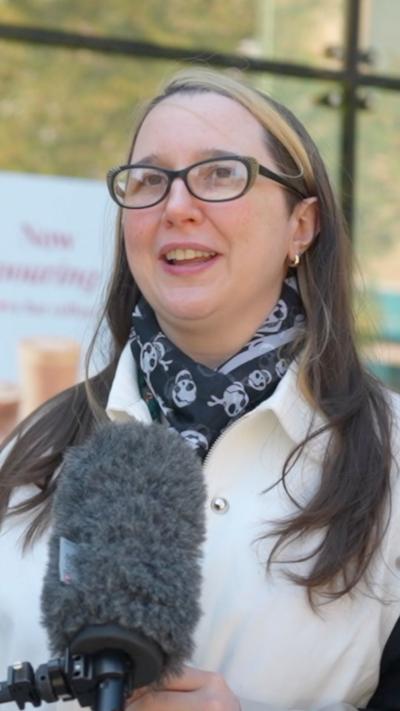 A woman is interviewed, with a microphone against blurred background of a sign and trees.