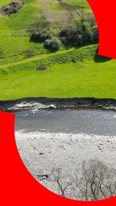A drone shot of the River Elwy, there is a completely dry grey riverbed next to the river which is now flowing into a nearby green field. In this bird's eye view shot there are also trees and a couple of small buildings. 