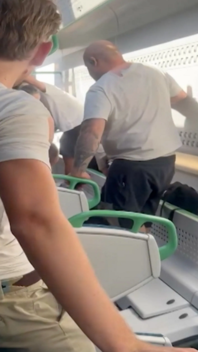 Video still taken from inside an overturned train carriage. Grey-backed seat with green handrails attached can be seen on their side as two men stand upright with their feet where carriage windows are