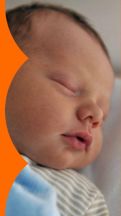A baby with a head of dark hair sleeps covered in a blue blanket
