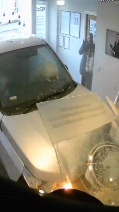 Silver car crashed into an office, with a shocked woman in the background.