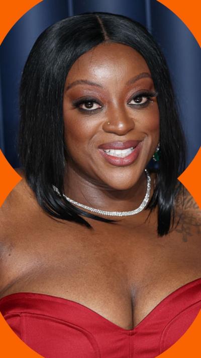 Judi Love smiles while wearing a red dress. She is wearing a silver necklace and has a tattoo on her left shoulder.