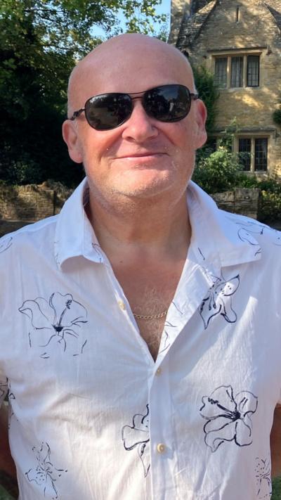A man in a white shirt and sunglasses smiles to the camera.