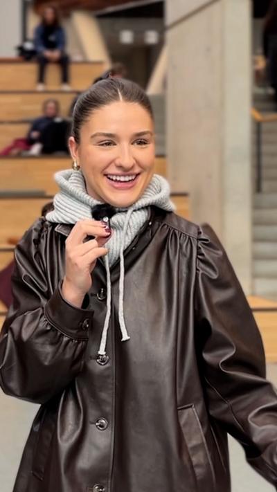 Student in a brown leather jacket with a grey hoody underneath. She has brown hair tied back and is holding a small microphone.
