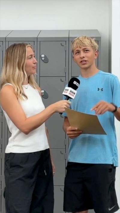 There are two people in frame standing in front of grey lockers, the person on the right hand side of the image is wearing a blue shirt and is holding a sand coloured envelope. The person on the left hand side of the image is wearing a white top and black bottoms, they are holding a microphone with bbc logos on it. Both people have blonde hair. 