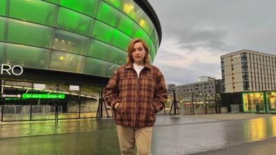 A girl with auburn hair is wearing a brown checkered coat with a white t-shirt underneath, light brown trousers and her hands are in her pocket. She is standing outside of the Ovo Hydro in Glasgow, which is lit up green. The sky is grey and the concrete ground looks wet and is reflecting the light.