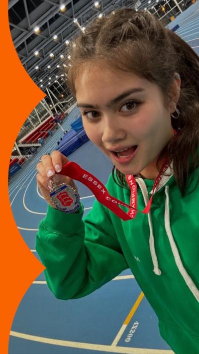 Athlete Issey Kyson holds up a medal while standing on an indoor race track. She has long brown hair and wears a green hoodie