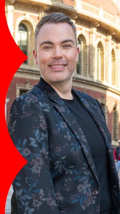 A man, Nicholas McCarthy, in a dark floral suit jacket stands in front of the Royal Albert Hall