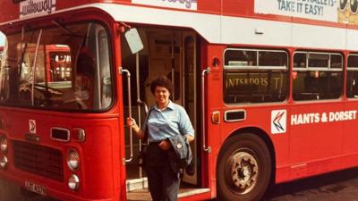 Gwen Watton wearing dark coloured trousers and a light blue shirt. She has short, curly dark hair and is standing beside a red double decker bus, resting her hand on its entrance railing.