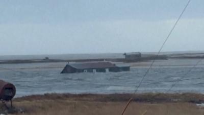 A house submerged almost up to its roof floats in water near land.