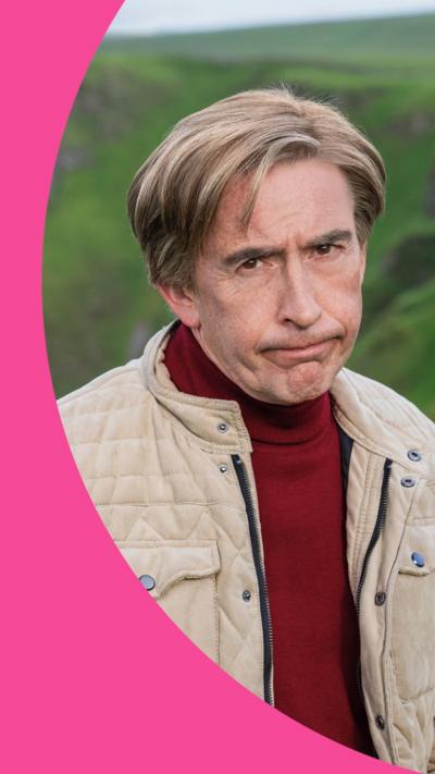 Alan Partridge stands in a field, looking unimpressed. He has dark blond hair and wears a beige jacket over a red jumper 