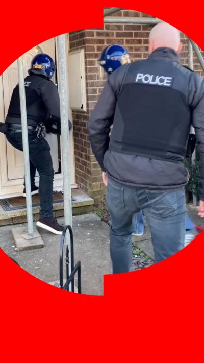 Two police officers in protective helmets break down a door, while a third watches.