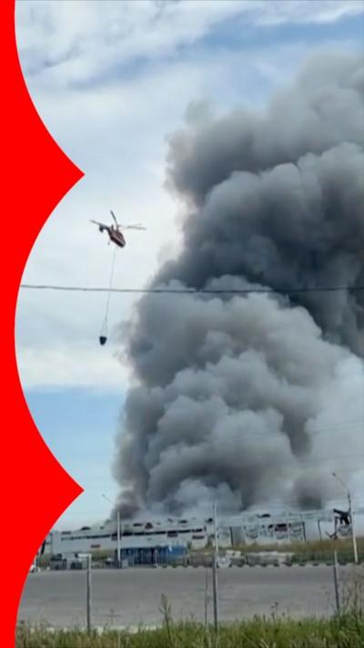 A helicopter carrying water flies towards a building with a huge plume of smoke erupting.