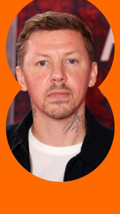 Professor Green has short blonde hair and is wearing a white t-shirt under a black jacket