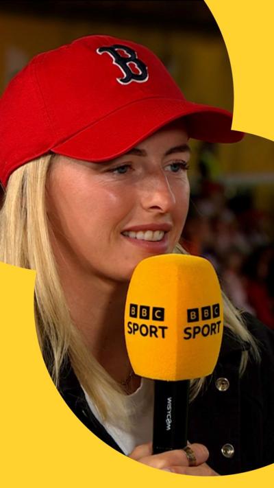 A blonde-haired woman wears a red baseball cap and holds yellow BBC Sport microphone