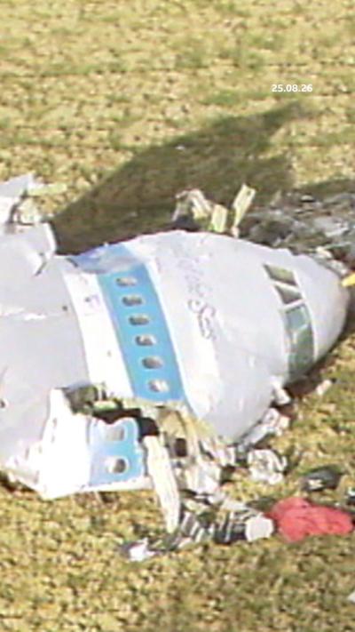 Archival image - a white plane with a light blue stripe, crashed in a field. Only the partial front end can be seen with debris strewn around.