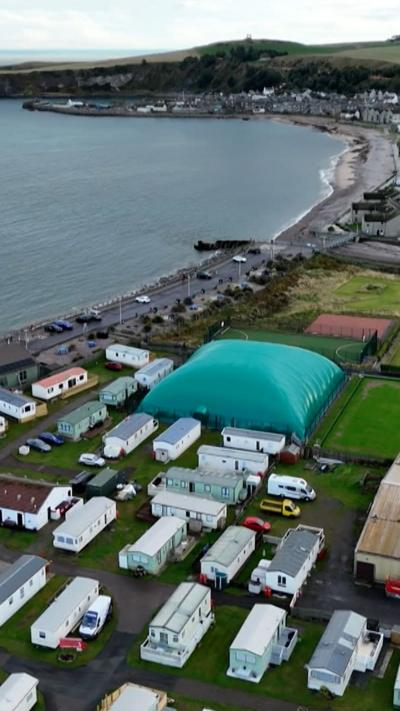 Aerial view of a giant inflatable green dome covering a tennis court beside a caravan park near the coast