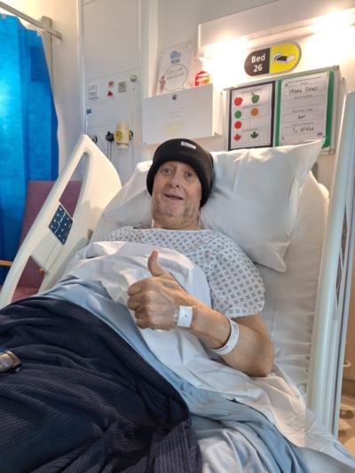 A man in a hospital bed wearing a black bobble hat giving the thumbs-up sign