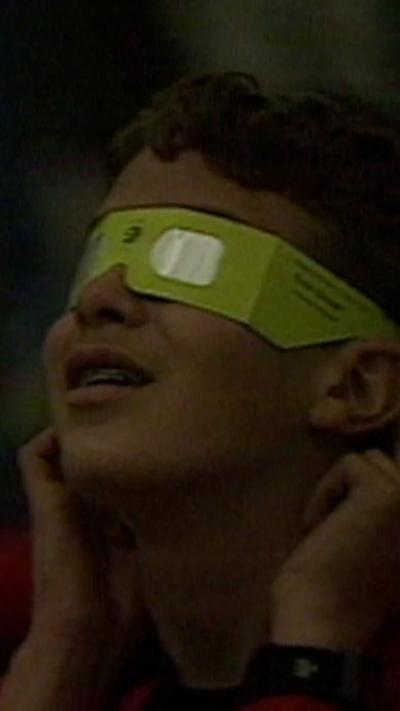 A boy wearing green eclipse glasses look up
