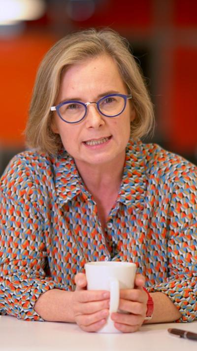 Felicity Evans on a table in a colourful patterned shirt. She is holding a white mug with both hands. She has blue framed circular glasses and short hair with a side parting 