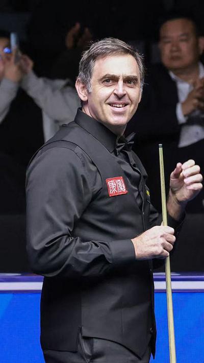 Ronnie O'Sullian hold his cue upright and smiles as he wears a black shirt and black waistcoat.