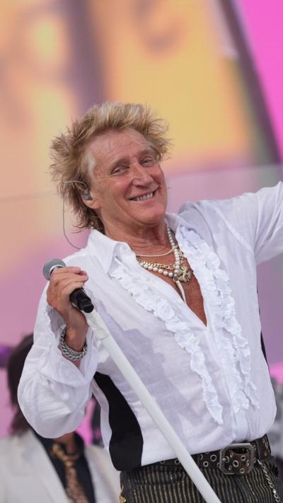 Rod Stewart wears an open neck white frilly shirt and holds a microphone, smiling at Glastonbury 2025