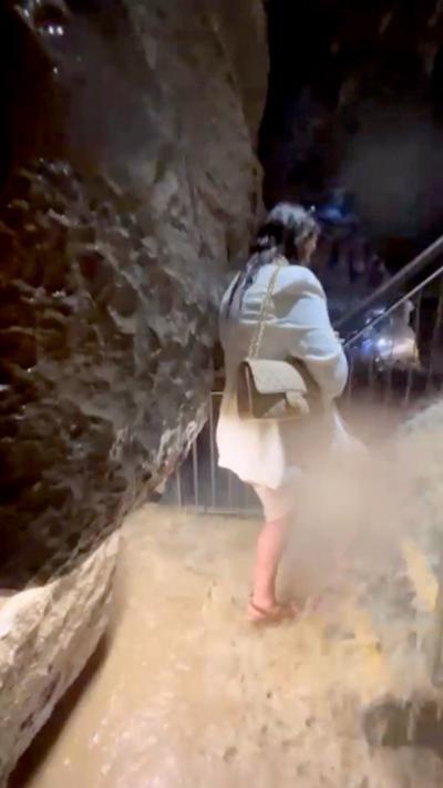 A woman in a cream coloured outside is walking up stairs inside a cave. There is a large volume of water flooding down the stairs.