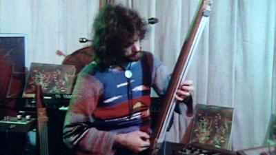 A man with long hair and a multicoloured jumper playing a kaleidophon.