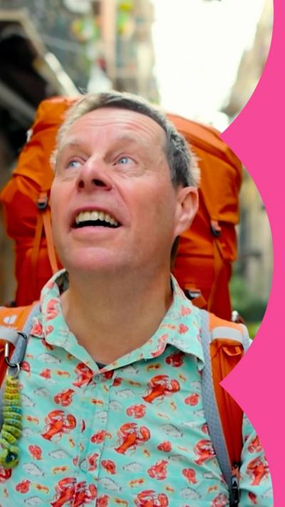 A man wearing a turquoise shirt with red lobsters on and an orange backpack looks up to the sky as he walks down a street.