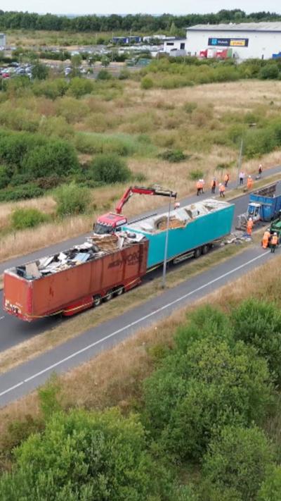 Drone shot of trailers dumped on road being emptied. The red and blue units have been left on a tarmac carriageway surrounded by grassed areas. Several workers wearing high visibility clothing can be seen cleaning the site.
