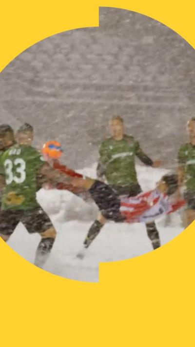 A group of footballers on a snow covered pitch. Some are wearing green shirts. One, in a red and white shirt, kicks an orange ball