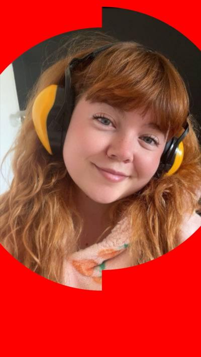 Celyn has long wavy red hair and wears ear defenders and a woolly peach top