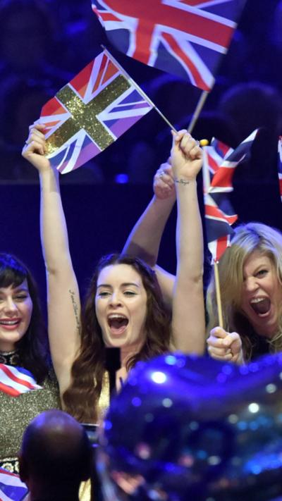 A woman with long dark hair and her arms in the air, a woman with long blonde hair. Both are holding small Union Jack flags on sticks and are screaming