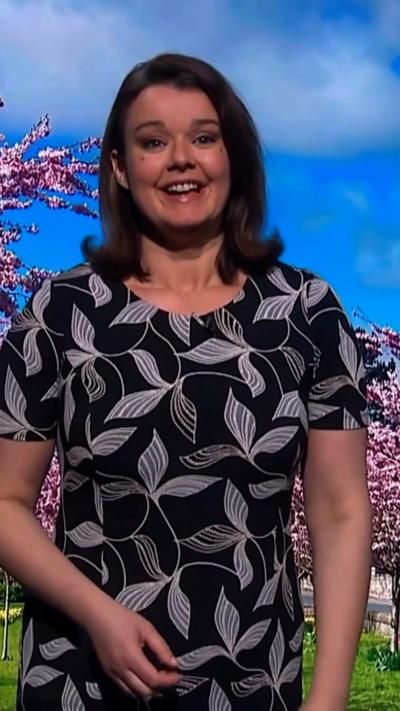Gillian Smart smiles as she stands in front of a green screen which is showing a blue sky, clouds, and pink trees. She has brown shoulder-length hair and is wearing a black dress with a white leaf design printed on it.