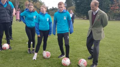 The Duke of Edinburgh is on the right wearing a tweed jacket, burgundy sweatshirt and grey trousers. He is kicking a football. Next to him on his right are three young people - a boy and two girls- wearing blue sports tops and black bottoms