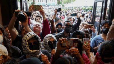 Protesters, some wearing face masks, sunglasses and head scarfs, enter an open door, with dozens more protesters behind them.