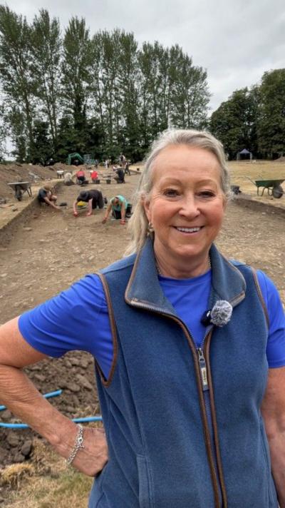 Melanie, wearing a blue top and fleece vest, stands in the foreground of an active archaeological excavation site, where several people are carefully digging and examining large rectangular trenches in a dry, open field bordered by trees.