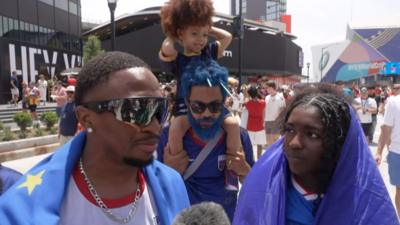 Four Cape Verde fans wearing blue and red, including a child sitting on a man's shoulders, stand to speak with a reporter.